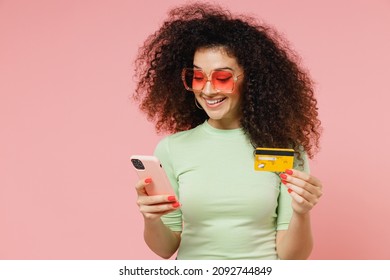 Young Curly Latin Woman 20s Years Old Wears Mint T-shirt Sunglasses Using Mobile Cell Phone Hold Credit Bank Card Doing Online Shopping Isolated On Plain Pastel Light Pink Background Studio Portrait