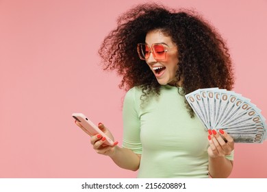 Young Curly Latin Woman 20s Years Old Wears Mint T-shirt Sunglasses Using Mobile Cell Phone Hold Fan Of Cash Money In Dollar Banknotes Isolated On Plain Pastel Light Pink Background Studio Portrait