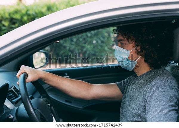 Young curly man driving a car in a medical mask\
and gray T-shirt. Man in protective mask is sitting at the wheel of\
a car. Protection from coronavirus epidemic pandemic, covid-19. New\
reality