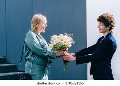 Young Curly Man Gives Flowers Older Woman. Smiling Adult Son Giving Flowers Daisies Bouquet To His Mother At City Street Outdoor.