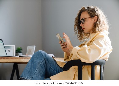 Young Curly Serious Woman In Glasses With Phone Sitting By Table In Cozy Room At Home