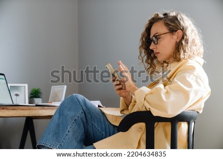 Young curly serious woman in glasses with phone sitting by table in cozy room at home