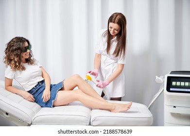 Young curly smiling woman in goggles on laser hair removal procedure in aesthetic clinic. Side view of process of laser hair removal on legs. - Shutterstock ID 2188578639