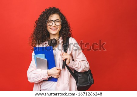 Young curly student woman wearing backpack glasses holding books and tablet over isolated red background. 