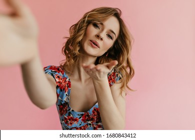 Young curly woman in bright floral outfit blows kiss on pink background. Stylish girl with blond hair takes selfie on isolated backdrop..