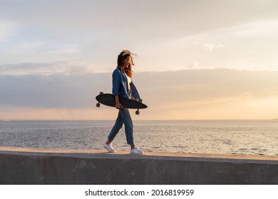 Young curly woman carry longboard at sunset looking at sea. Stylish caucasian skateboarder female enjoy evening sunlight at seaside walking at pavement. Freedom, street fashion and lifestyle concept - Powered by Shutterstock