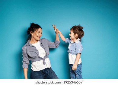 Young curly woman and cute little girl look at each other and clapping hands gesturing "high five". Mother and and daughter on blue background - Powered by Shutterstock