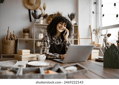 Young curly woman potter taking order for purchase of tableware using phone and laptop. Happy female with new purchase in ceramics shop sitting at table against background of shelves with dishes - Powered by Shutterstock