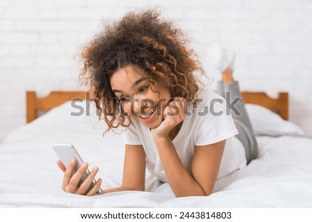 A young curly-haired african american woman intently gazes at her mobile phone while propping herself up in bed with a contemplative expression