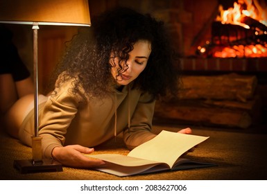 A Young Curly-haired Lady In A Panties Reads A Book, Lying On The Floor With A Cozy And Warm Fireplace In The Background. Piermont Retreat, Tasmania, Australia. Portrait Photography.