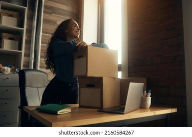 A young curly-haired logistics employee packs goods, collects parcels, puts things in a cardboard box, prepares a parcel for shipment, transports, sits on a windowsill and drinks a report in a book - Powered by Shutterstock