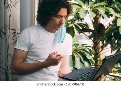 Young curly-haired man removes a medical mask from his face in a white T-shirt with a black laptop near the window. He reads news about the end of quarantine from the coronavirus. Waiting for work - Powered by Shutterstock