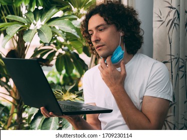 Young curly-haired man removes a medical mask from his face in a white T-shirt with a black laptop near the window. He reads news about the end of quarantine from the coronavirus. Waiting for work - Powered by Shutterstock