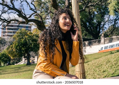 Young Curly-haired Woman Happily Talking On The Cell Phone In A Public Square. Mobile Techonology, 5g, Communications Concepts.