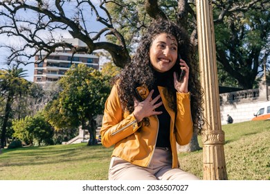 Young Curly-haired Woman Happily Talking On The Cell Phone In A Public Square. Mobile Techonology, 5g, Communications Concepts.