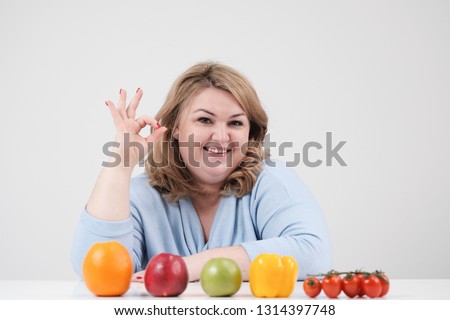 Young curvy fat woman in casual blue clothes on a white background at the table and shows OK, vegetables and fruits are laid out in front of her in a row. Diet and proper nutrition, and harmful food.