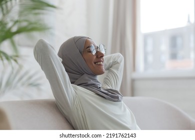 Young curvy muslim woman in hijab and glasses resting at home, woman on sofa with arms behind head, smiling with closed eyes - Powered by Shutterstock