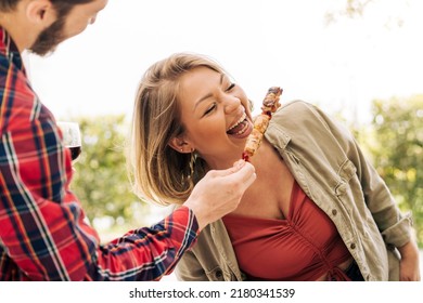 A young curvy woman laughs as she eats skewers of meat from her friend's hands during a trip to the countryside. Young adults people having fun together at picnic - people lifestyle concept