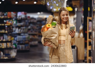 Young customer woman wear casual clothes hold paper bag use mobile cell phone shopping at supermaket store grocery shop buying choose products inside hypermarket. Purchasing food gastronomy concept - Powered by Shutterstock