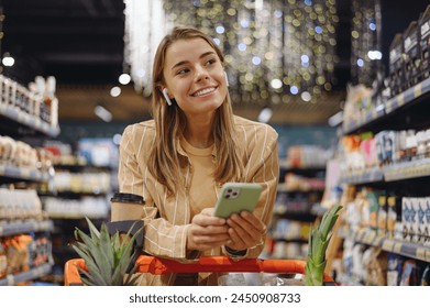 Young customer woman wear casual clothes earphones listen music use mobile cell phone shopping at supermaket store grocery shop buy with cart choose products in hypermarket. Purchasing food concept - Powered by Shutterstock