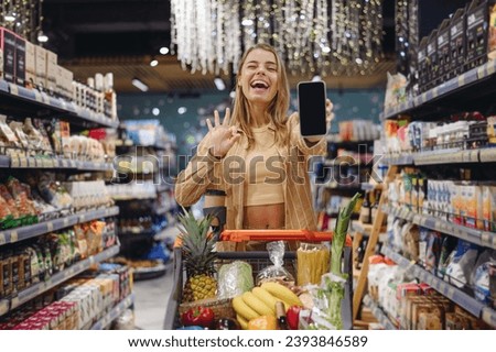 Young customer woman wear casual clothes showing blank screen mobile cell phone shopping at supermaket store grocery shop buying with trolley cart choose product in hypermarket Purchasing food concept