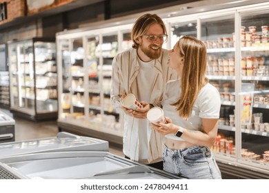 Young customers laughing and buying groceries, choosing a product from a shelf in the frozen food section at the supermarket - Powered by Shutterstock