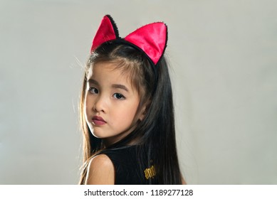 Young Cute Asian Beautiful Eyes Girl Kid Dressing In Black Devil Fashion Costume With Cat Ears Portrait Photo Shooting In Studio. Seen From Side Facing. Devil Fashion And Halloween Portrait Concept.