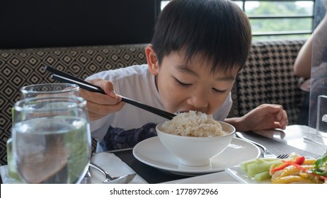 The Young Cute Asian Boy Is Eating Hungrily A Big Bowl Of Rice By Using Chopsticks At The Dining Table.