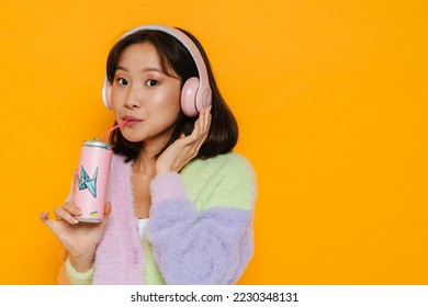 Young cute asian woman in headphones drinking soda isolated over yellow studio background - Powered by Shutterstock