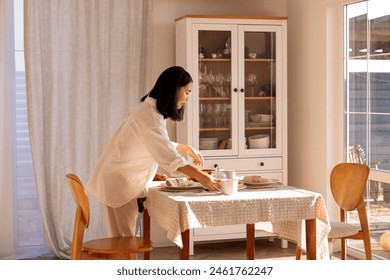 A young cute Asian woman sets the table in the dining room at home. A charming Korean girl sets out plates and cups for a family breakfast. Household chores and routine. - Powered by Shutterstock