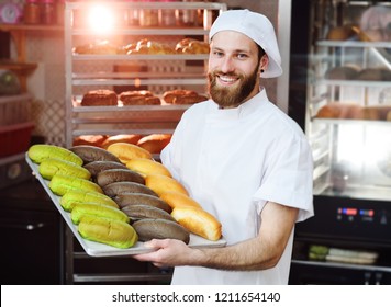 Young Cute Baker In White Uniform Holding A Tray With Colored Rolls For Hot Dog On The Background Of Bakery Or Bread Factory