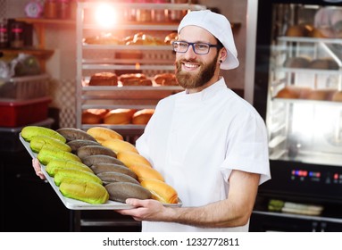 Young Cute Baker In White Uniform Holding A Tray With Colored Rolls For Hot Dog On The Background Of Bakery Or Bread Factory