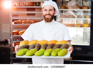 Young Cute Baker In White Uniform Holding A Tray With Colored Rolls For Hot Dog On The Background Of Bakery Or Bread Factory