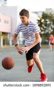 Young Cute Boy Plays Basketball At The Outdoor Streetball Court On A Sunny Summer Day. Teenager Player In Action Dribbling The Ball. Hobby, Active Lifestyle, Sports For Kids.