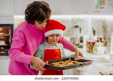 Young Cute Boy In Winter Shirt And Red Santa's Hat Is Cooking Christmas Cookies On White Home Kitchen With His Attractive Grandmother. Holiday Concept.