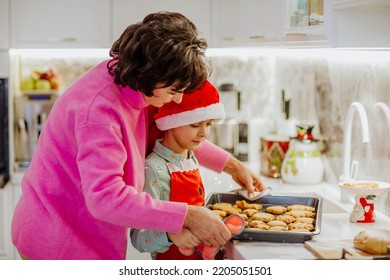 Young Cute Boy In Winter Shirt And Red Santa's Hat Is Cooking Christmas Cookies On White Home Kitchen With His Attractive Grandmother. Holiday Concept.