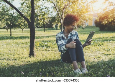 Young Cute Brazilian Girl With Digital Tablet Sitting On Grass Of City Park And Reading Electronic EBook, Beautiful Mixed Female Teen With Afro Hair, Springtime Meadow With Copy Space For Your Message