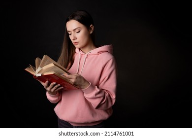 A Young Cute Brunette In A Pink Hoodie, Reading A Book. Student With An Open Book In Her Hands, Portrait On Black. Study Or Read Fiction