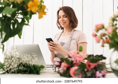 Young cute cheerful florist lady standing with flowers in workshop using laptop computer chatting by mobile phone. - Powered by Shutterstock