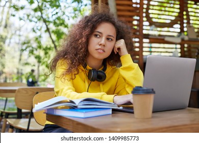 Young Cute Dark Skinned Curly Girl In Yellow Sweatshirt, Sits On A Cafe Terrace, Preparing For Exam On A Laptop, Looks Thoughtfully Into The Distance, Thinks Which Topic Is Best To Write An Essay.