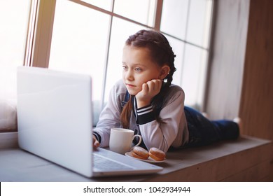 Young cute emotional teenager girl with braids in overalls lies by the window indoor. With cup and cookies, working with the laptop - Shutterstock ID 1042934944