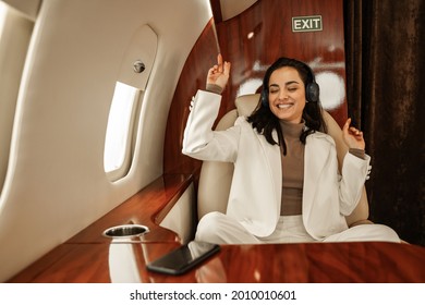 Young Cute Girl Airplane Passenger Having Fun With Incendiary Music Via Bluetooth Headphones Connected To Her Phone Sitting On A Leather Chair VIP Private Business Jet