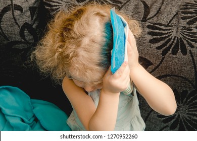 Young Cute Girl Sit On Couch In Home Living Room And Holds Cold Gel Ice Pack On Head. Headache, Fever Or Concussion Concept.
