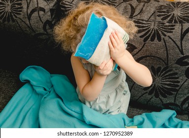 Young Cute Girl Sit On Couch In Home Living Room And Holds Cold Gel Ice Pack On Head. Headache, Fever Or Concussion Concept.