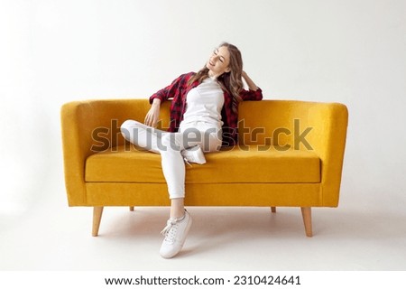 young cute girl sits on soft comfortable sofa and relaxes, pensive woman is resting on yellow couch on white isolated background