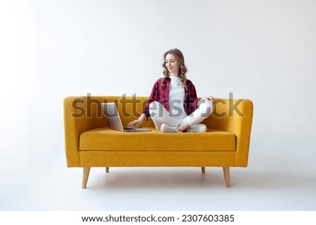 young cute girl uses laptop on comfortable soft sofa, woman types online on computer on yellow couch on white isolated background