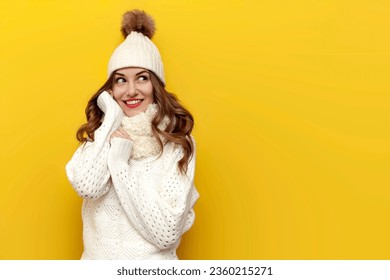 young cute girl in warm soft winter clothes smiles on yellow isolated background, woman in white hat and scarf touches comfortable and cozy knitted sweater