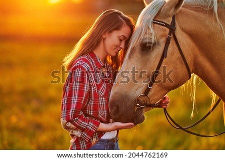 Young cute happy joyful satisfied smiling woman with closed eyes enjoying of hugging and stroking beautiful blond palomino horse at meadow at sunset
