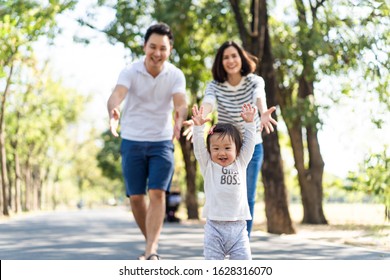 Young Cute Happy Little Asian Toddler Girl Running In Park With Parents. Mom Dad Taking Care Daughter By Following Looking After Carefully. Kid Smiling Enjoy Learning To Walk And Run, Family Relation.