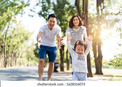 Young Cute Happy Little Asian Toddler Girl Running In Park With Parents. Mom Dad Taking Care Daughter By Following Looking After Carefully. Kid Smiling Enjoy Learning To Walk And Run, Family Relation.